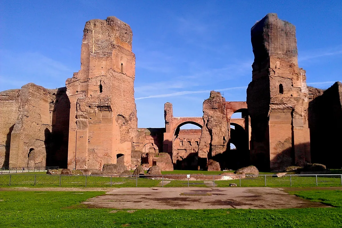 The tall brick ruins of the Baths of Caracalla in Rome against a blue sky, with a grassy lawn in front