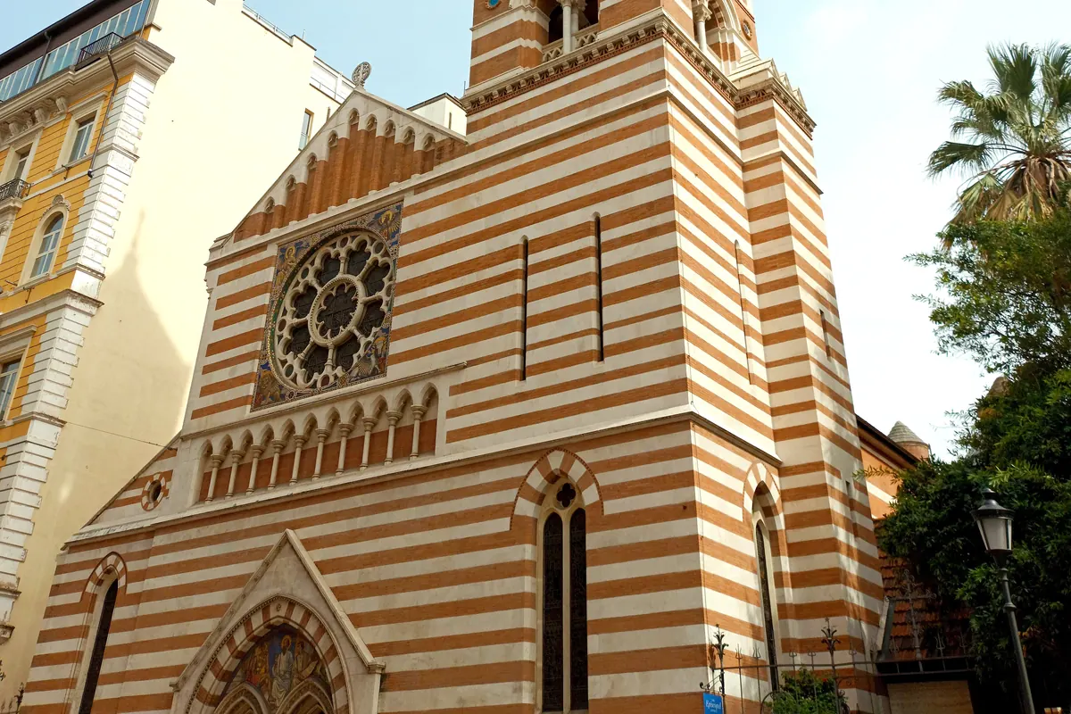 The front of a church in horizontal bands of red brick and white stone, with a carved rose window framed by mosaic, an arcade of small arches and a Gothic portal.